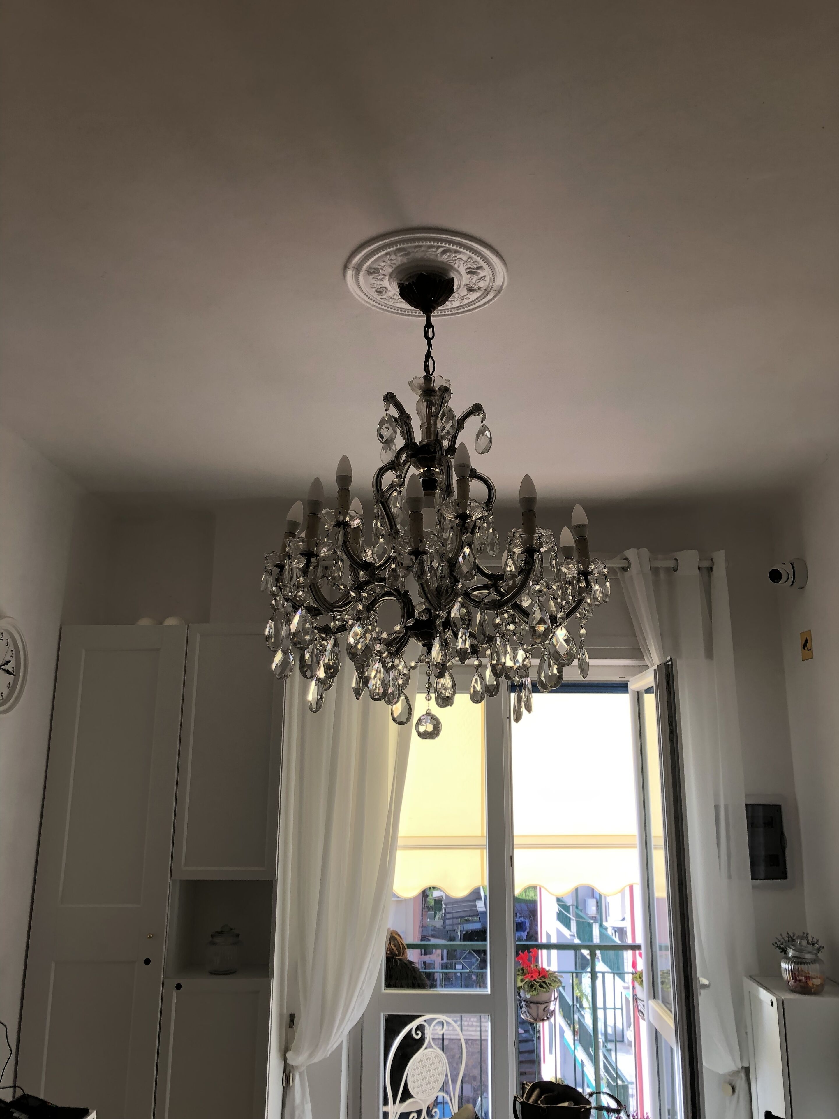 A room with a chandelier, white curtains, a window with a view of buildings, and a clock on the wall.