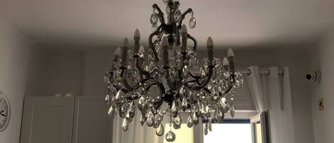 A room with a chandelier, white curtains, a window with a view of buildings, and a clock on the wall.