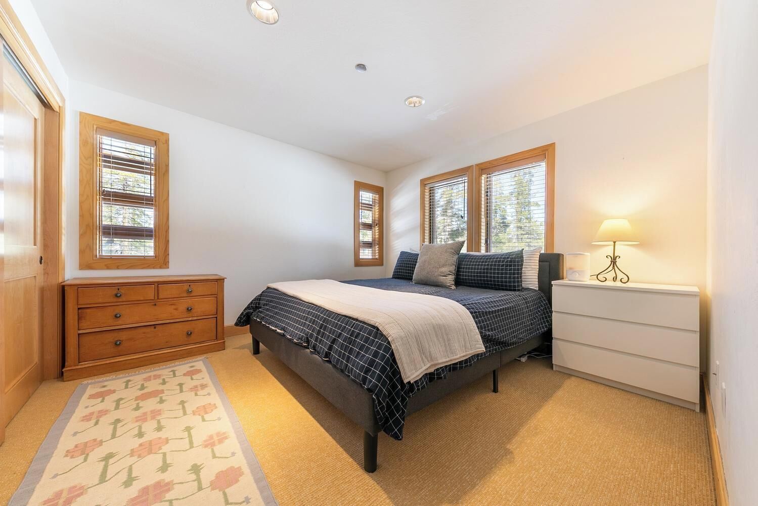 Une chambre à coucher avec un lit, une commode, une table de chevet, une lampe et des fenêtres.