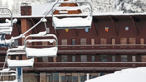 Un asiento de telesilla cubierto de nieve, un edificio de madera con varias ventanas y una montaña nevada al fondo.