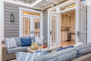 A covered patio area with a wicker sofa, a table with a basket of fruit and bottles, and a kitchen visible through glass doors. - Stunning 5bdr Minutes to Beach, Pier, Restaurants Emerald Cove by Avantstay (Newport Beach)
