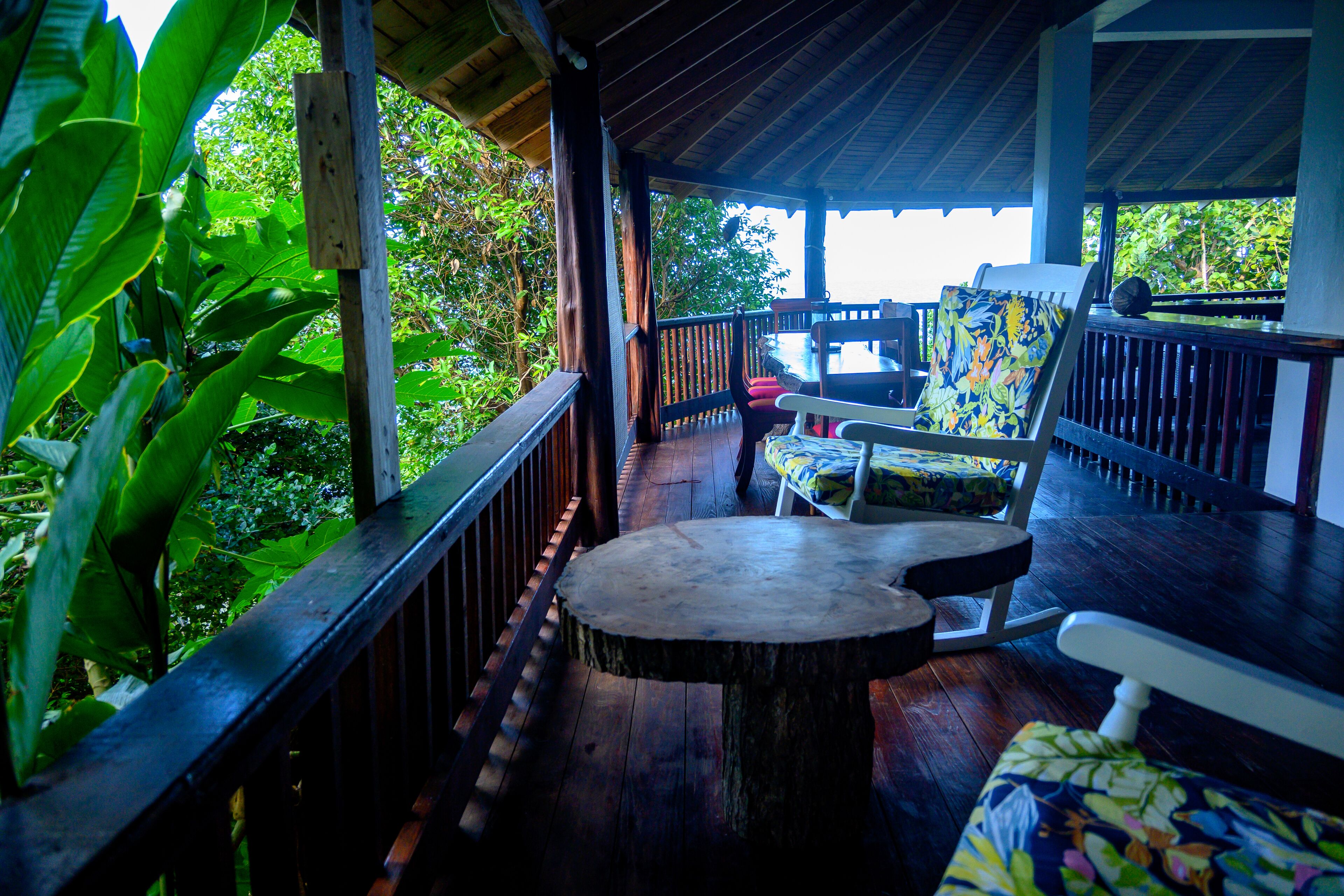 A wooden deck with a round table and two chairs, surrounded by lush greenery.