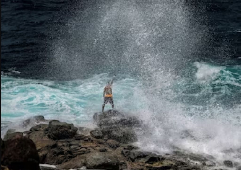 Una persona de pie sobre rocas junto al mar, con olas rompiendo a su alrededor.