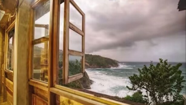 A room with a large window overlooking a coastal view with waves and a hilly landscape. - Private wooden villa with stunning ocean views, in an enchanting garden setting. (Salybia Kalinago Territory)