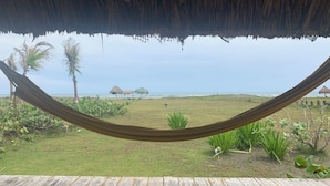 Un'amaca sotto un tetto di paglia, con vista su una zona erbosa, palme e una spiaggia in lontananza.
