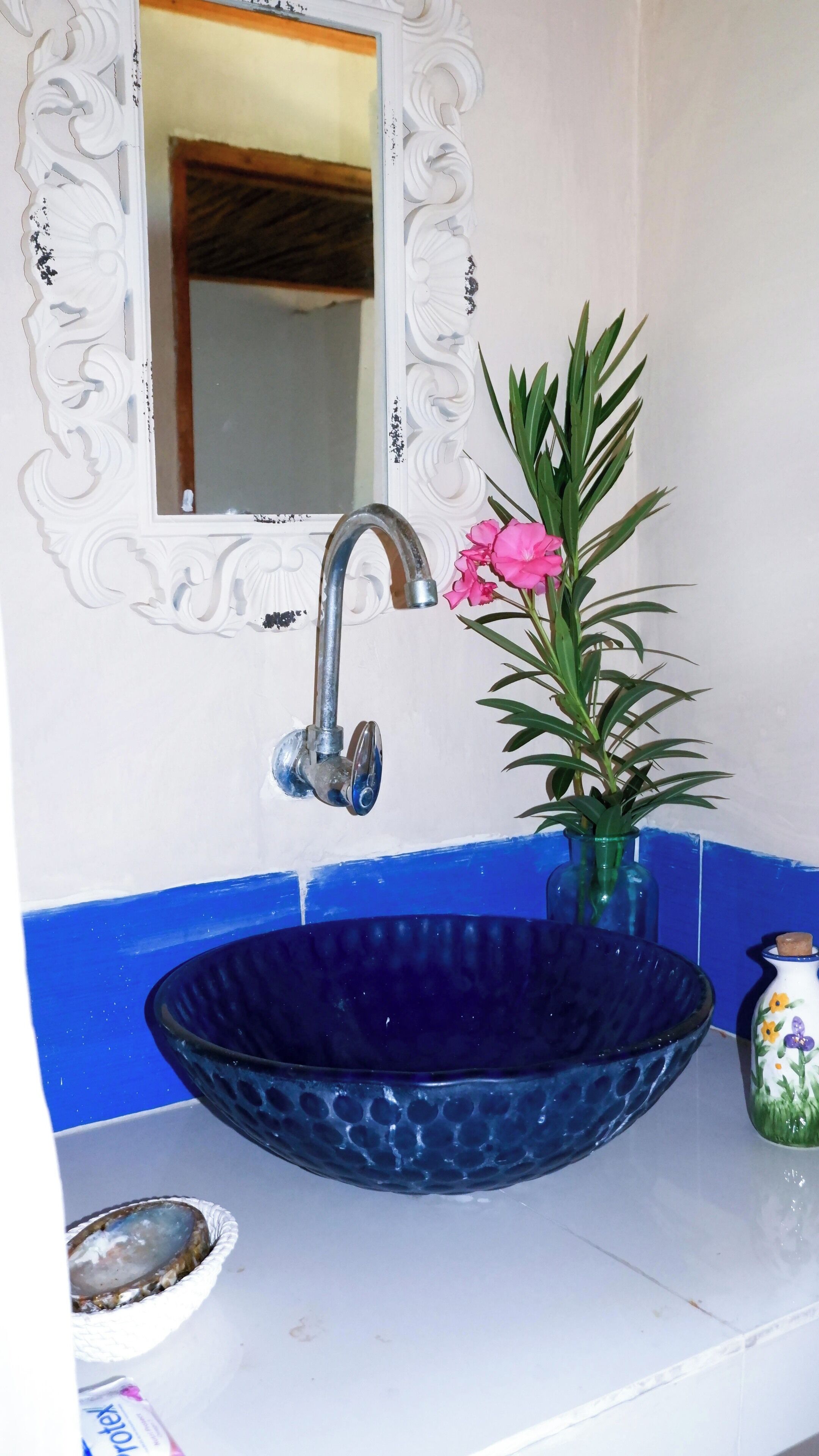 A bathroom with a blue sink, a white mirror with decorative molding, a potted plant with pink flowers, and a small decorative vase.