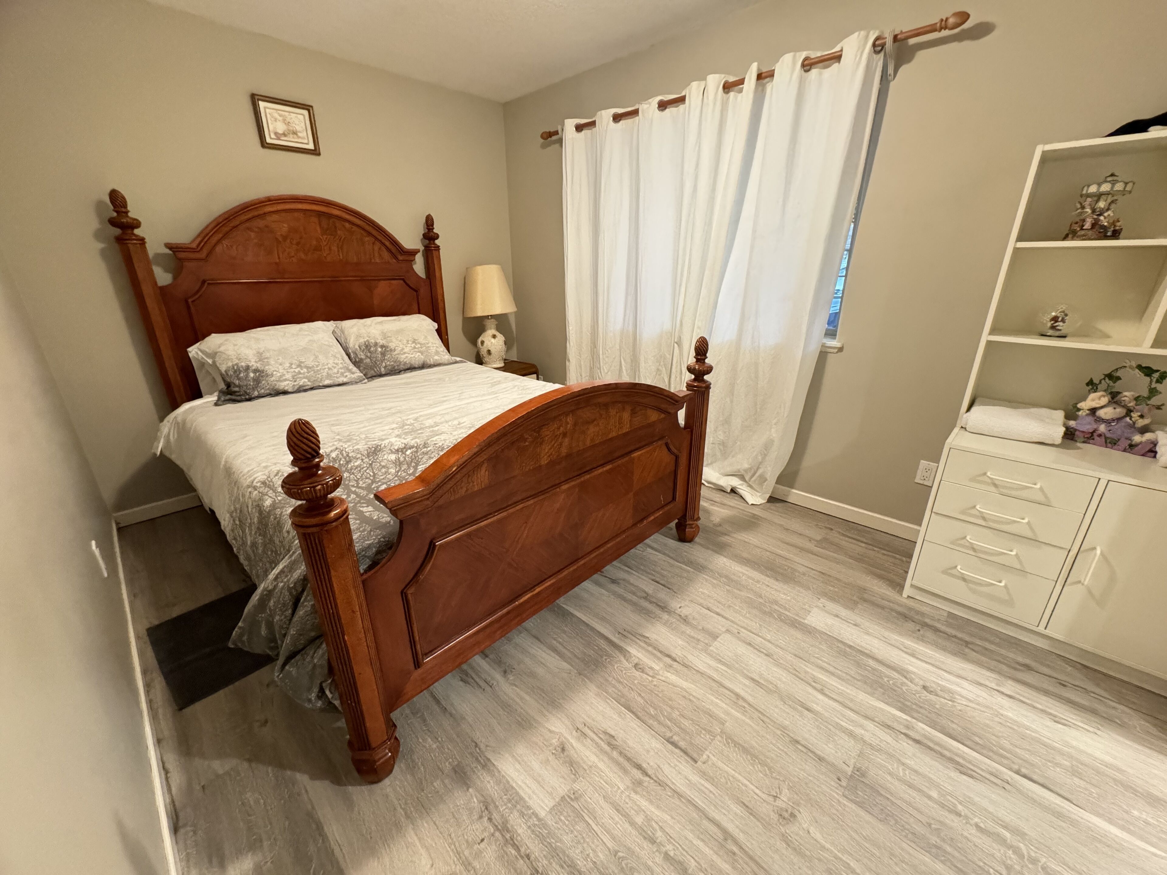 A bedroom with a wooden bed, a nightstand, a lamp, a white dresser, and a window with curtains.