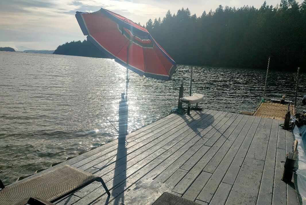 Um cais de madeira com um guarda-sol vermelho, um banco e vista para um lago calmo com uma ilha arborizada ao longe.
