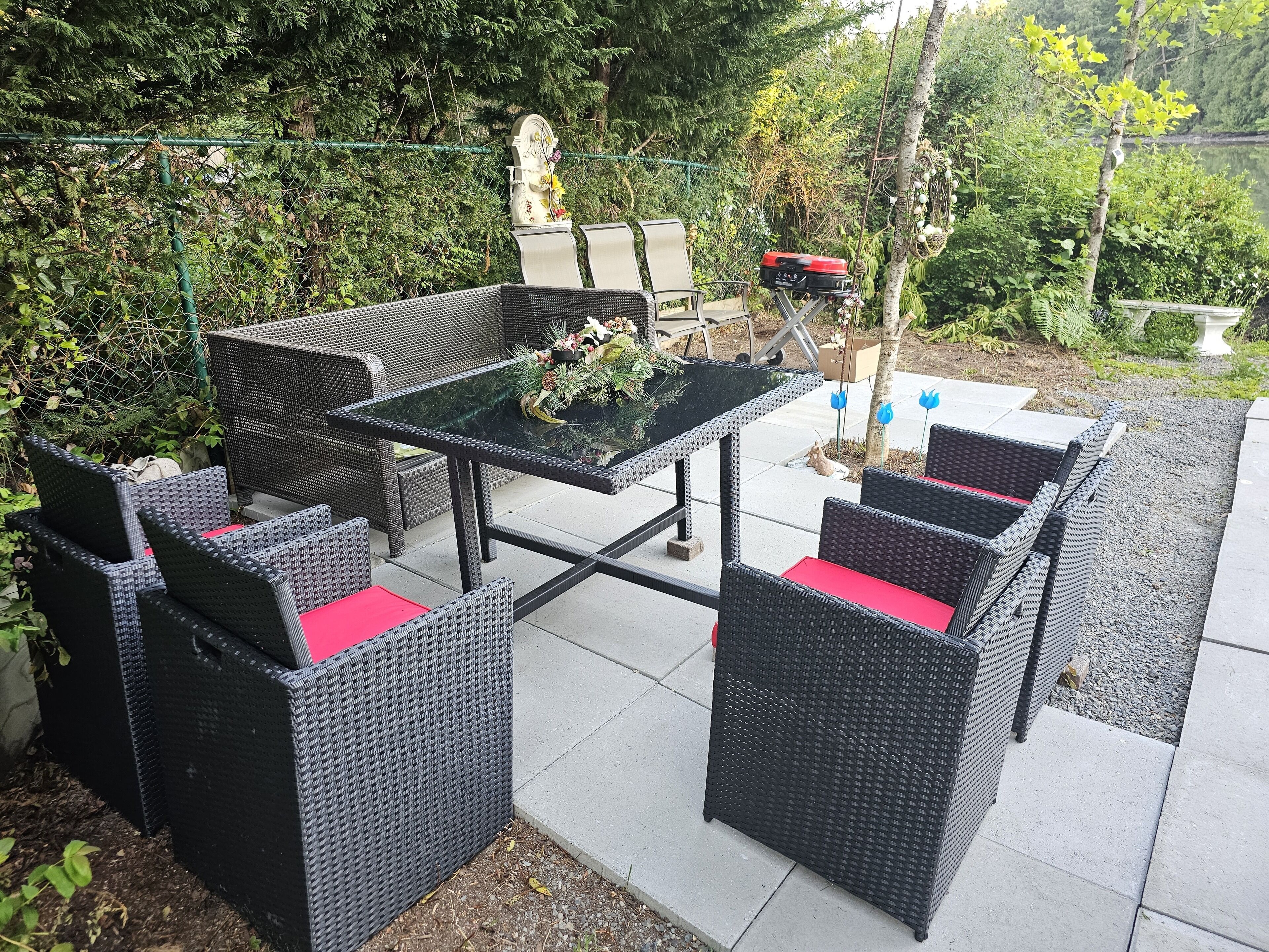 An outdoor patio with wicker furniture and a glass table, surrounded by greenery and a fence.