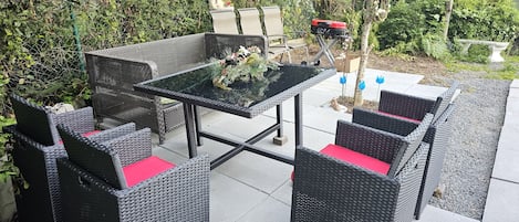 Un patio al aire libre con mobiliario de mimbre y una mesa de vidrio, rodeado de follaje y una cerca.