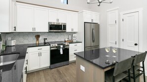 A modern kitchen with white cabinets, stainless steel appliances, a black countertop, and a dining area with two chairs. - Rise & Wine | Hot Tub | Sleeps 4 (Fredericksburg)