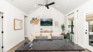 A modern kitchen with a large island, a ceiling fan, and a dining area with a sofa and table. - Rise & Wine | Hot Tub | Sleeps 4 (Fredericksburg)