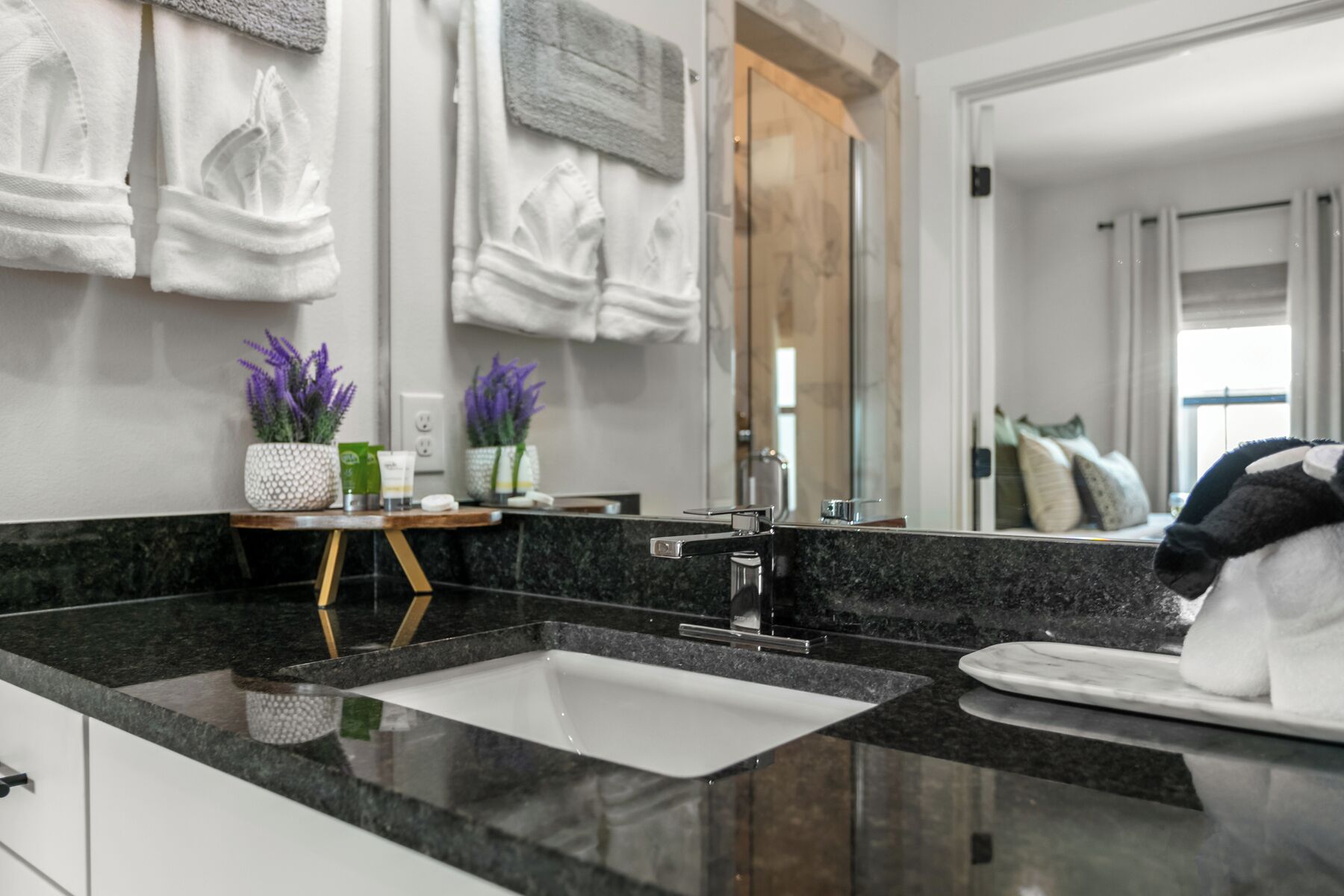A modern bathroom with a black granite countertop, a white sink, and a large mirror. There are towels hanging on the wall and a small wooden stool with potted lavender plants.