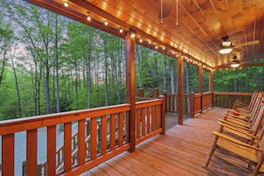 A wooden deck with a railing, surrounded by trees, and furnished with wooden chairs.
