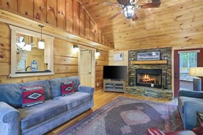 A wooden cabin interior with a sofa, television, fireplace, and ceiling fan. - Upscale Morganton Cabin w/ Hot Tub & Arcade (Morganton)