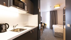 Une chambre d’hôtel de taille réduite, équipée d’une kitchenette, d’un lit, d’un bureau et d’une salle de bain.