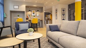 Un moderno vestíbulo de hotel con un sofá gris, una silla amarilla y una mesa redonda con una planta. Hay cuadros enmarcados en las paredes y una planta pequeña en un estante.
