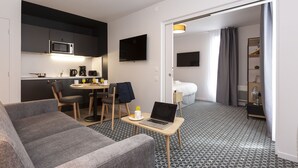 Una habitación de hotel moderna con kitchenette, zona de comedor, sofá y un portátil sobre una mesita.