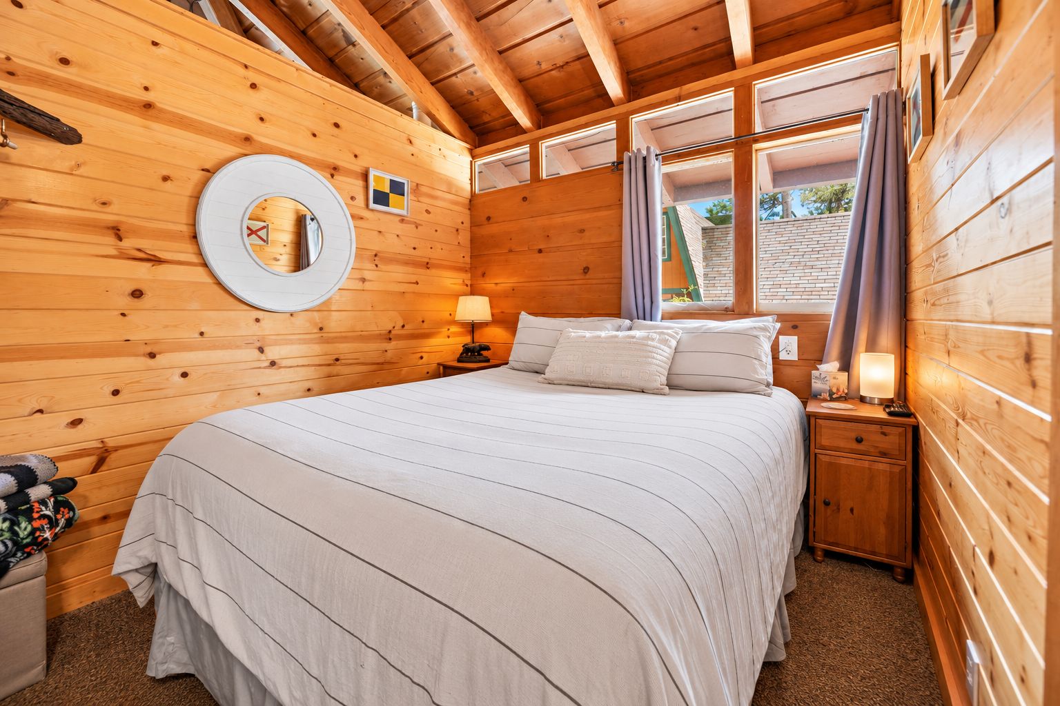 A wooden cabin bedroom with a bed, nightstands, a lamp, a mirror, and a window with curtains.