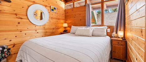 A wooden cabin bedroom with a bed, nightstands, a lamp, a mirror, and a window with curtains.