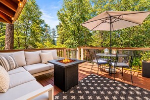 An outdoor deck with a sofa, a fire pit, a table with chairs, and an umbrella. - Clerestory Cottage | Private Hot Tub, Sauna & Walk to Big Bear Lake (Big Bear Lake)