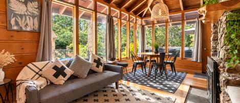 A cozy room with a sofa, dining table, and chairs, large windows, and a stone wall.