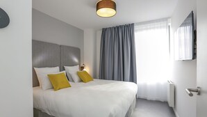 Ein modernes Hotelzimmer mit einem großen Bett, einem Kopfteil, einem Nachttisch mit Lampe, einem Fenster mit Vorhängen und einem an der Wand montierten Fernseher.