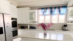 A modern kitchen with white cabinetry, a central island, stainless steel appliances, and a window with blue curtains. - Home Away From home. (Bridgetown)