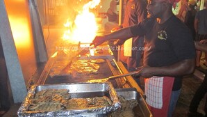A person grilling meat on a large outdoor grill with flames visible. - Home Away From home. (Bridgetown)