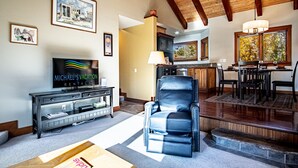 Een woonkamer met een televisie, een lederen fauteuil en een eethoek met een tafel en stoelen.