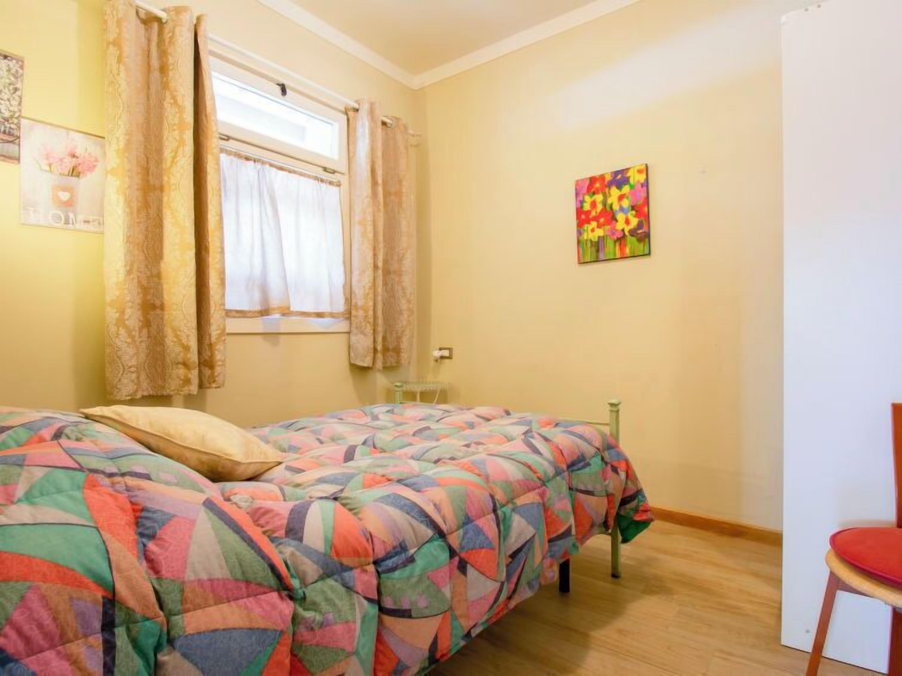 Een slaapkamer met een bed, gordijnen, een raam, een stoel en een kleurrijk schilderij aan de muur.