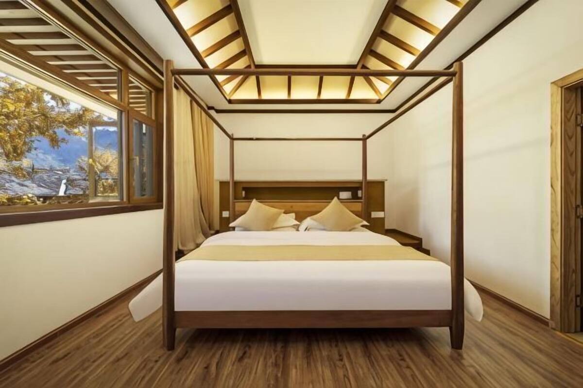 A four-poster bed with a wooden frame and white bedding, a wooden floor, and a window with a view of trees.