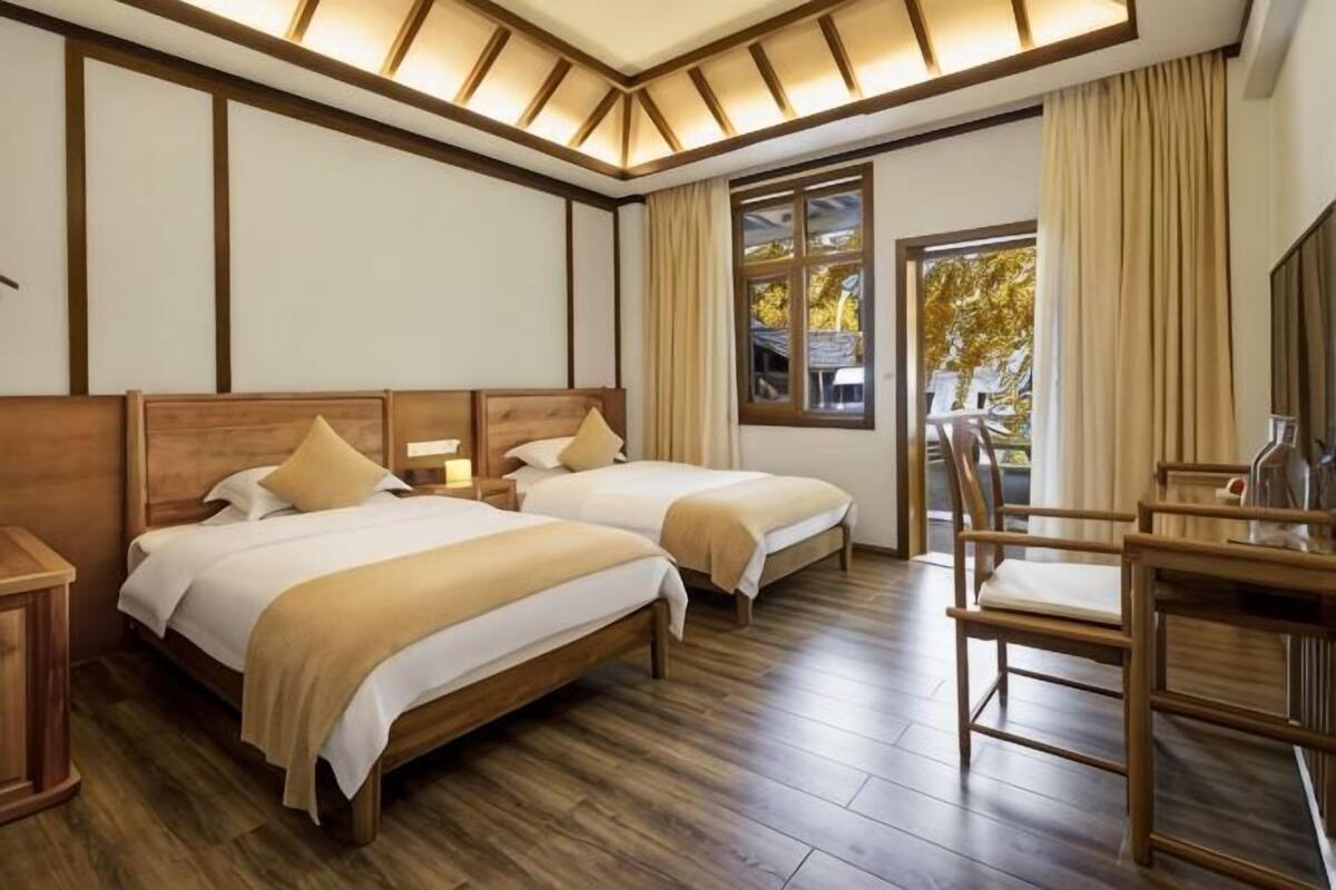 A hotel room with two beds, wooden flooring, a desk, and a window with curtains.