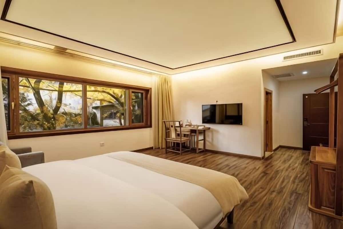 A bedroom with a large bed, a desk, a chair, a TV, and a window with a view of trees.