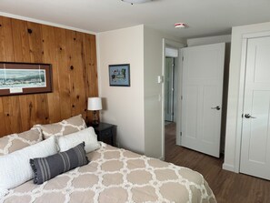 Un dormitorio con pared de paneles de madera, una cama con ropa de cama estampada, una mesita de noche con una lámpara y una puerta que da a otra habitación.