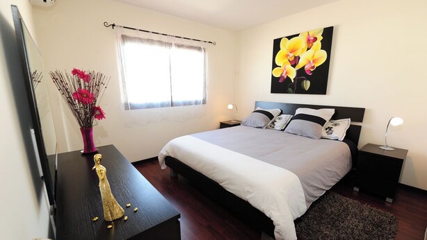 Un dormitorio con cama, mesitas de noche, cómoda, espejo, ventana con cortinas y un cuadro en la pared.