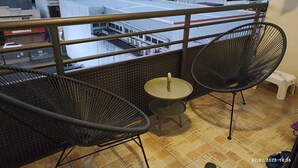 Un balcón con dos sillas de mimbre negras y una mesita redonda, suelo de baldosas y vistas a edificios al fondo.
