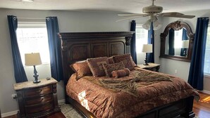 A bedroom with a large bed, wooden headboard, two bedside tables with lamps, a ceiling fan, and a large mirror. - The Whitehouse. (Green Cove Springs)