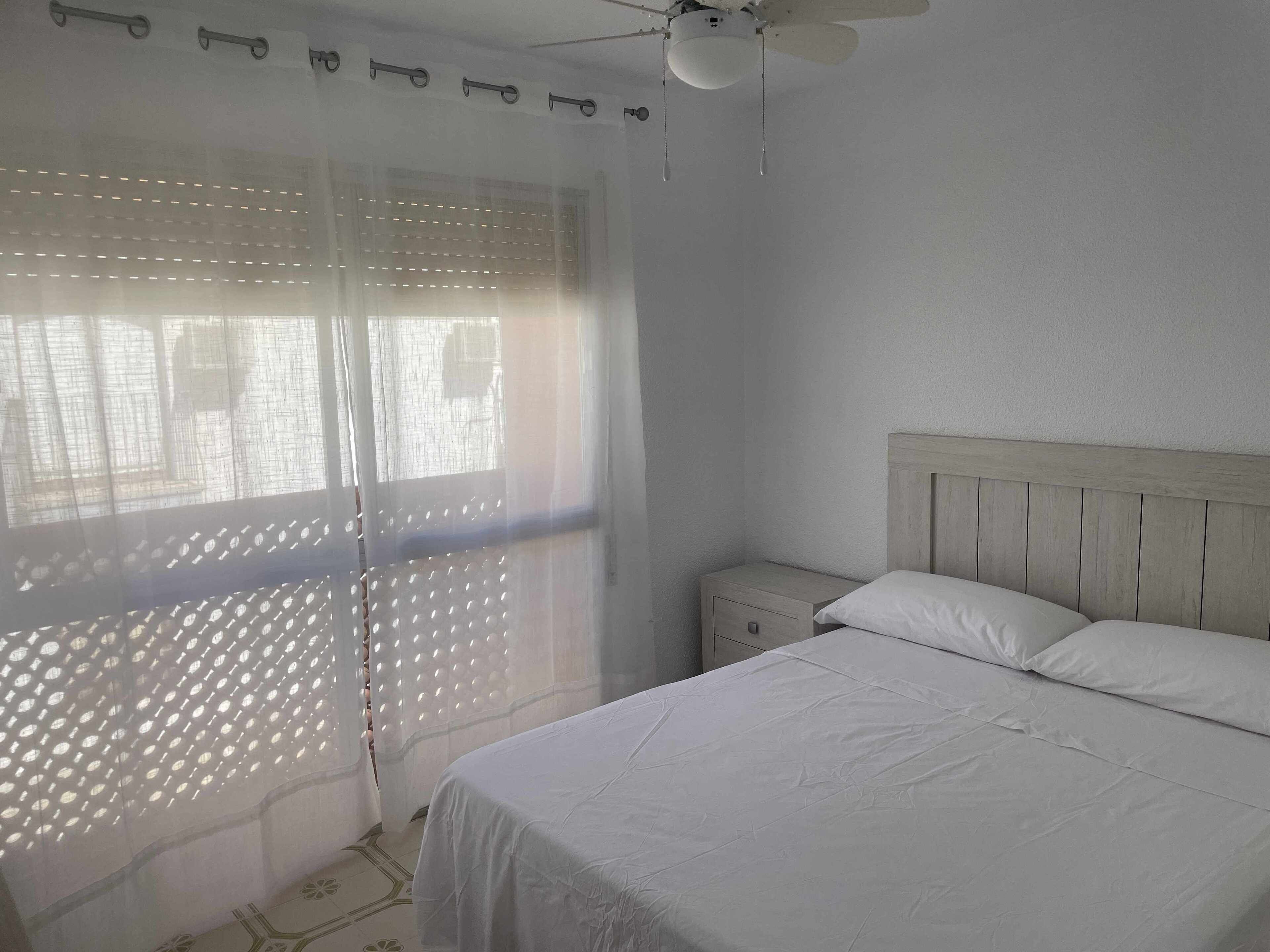 A bedroom with a bed, a nightstand, a ceiling fan, and a window with sheer curtains.