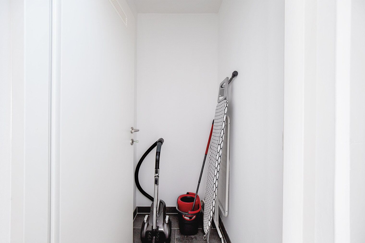A small, narrow room with a door, a vacuum cleaner, a red bucket, and a pair of skis leaning against the wall.