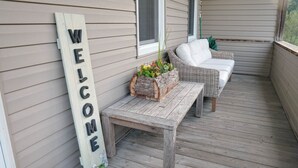 Eine Holzterrasse mit einer Rattan-Couch und einer Bank, einem Willkommensschild und einem Blumenkasten mit Blumen.