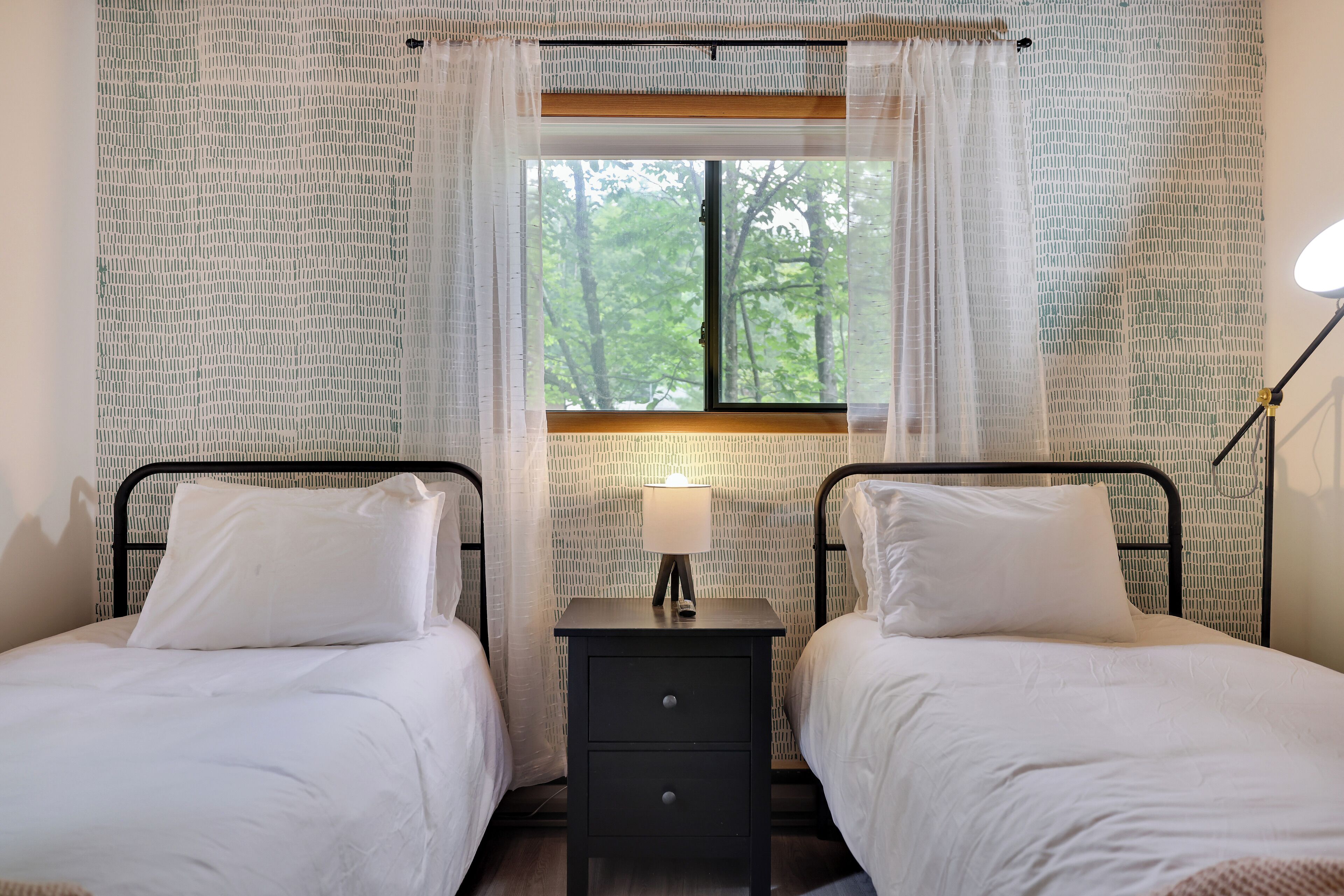 A bedroom with two beds, a nightstand with a lamp, and a window with a view of trees.