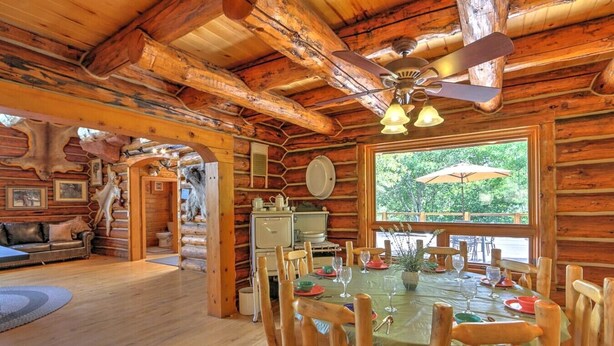 A rustic wooden cabin interior with a dining table set for four, a television, and a large window with an umbrella outside.