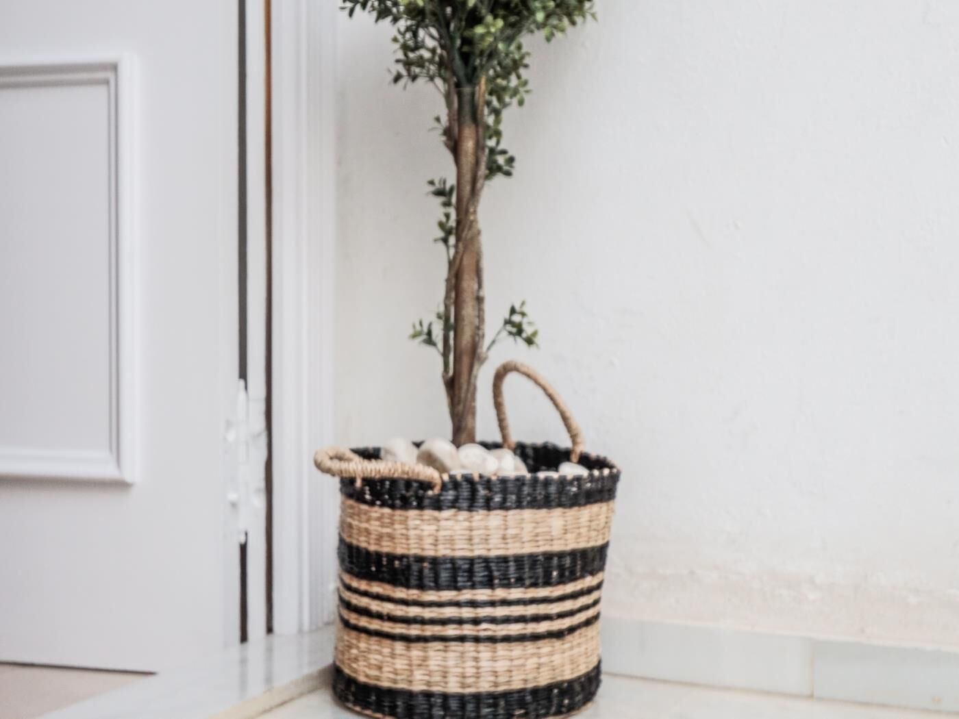 A woven basket with handles, placed on a tiled floor next to a white door.