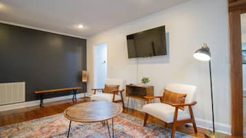 Una sala de estar con una mesa redonda, dos sillones blancos, un banco y una lámpara de pie.