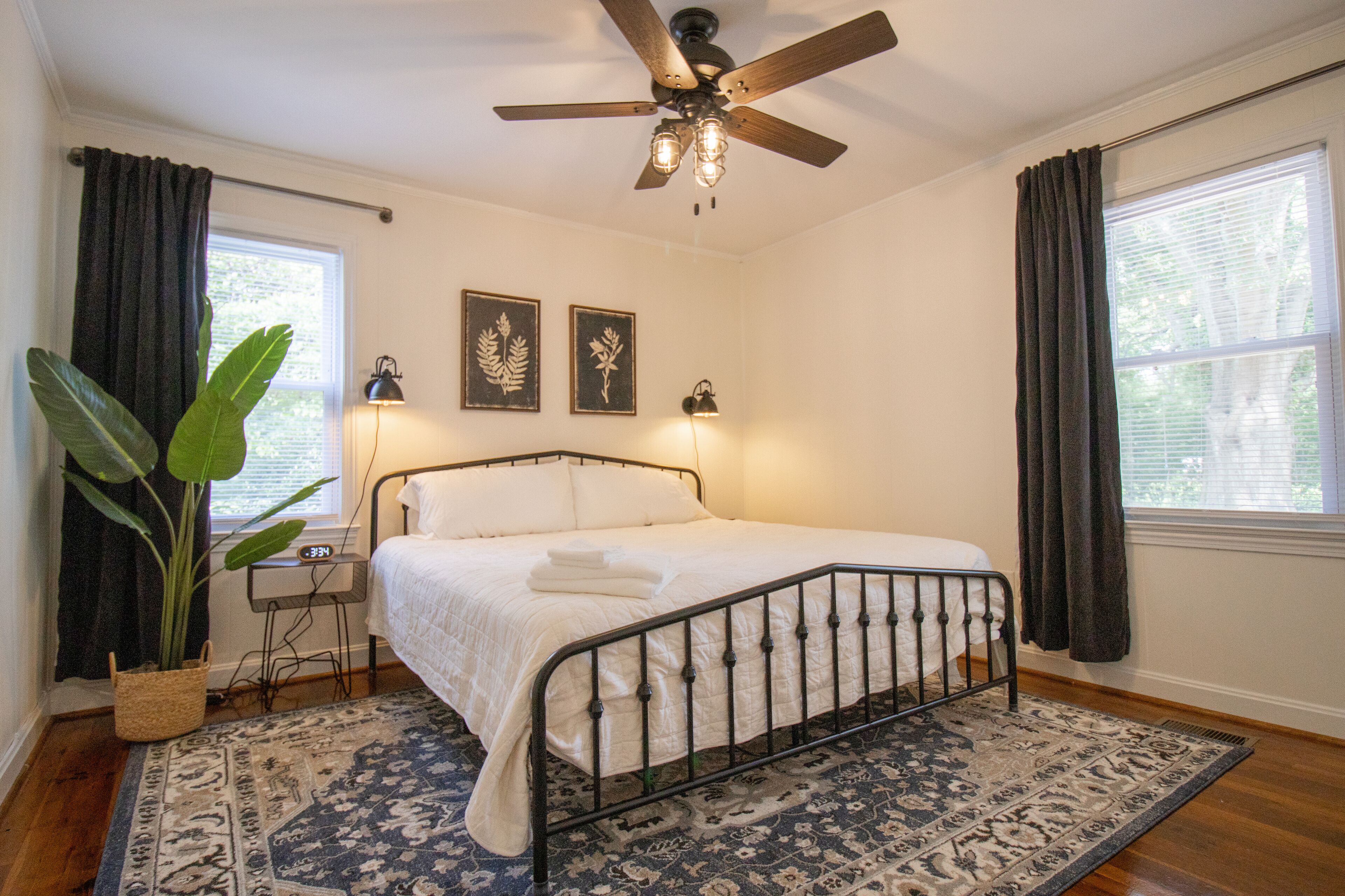 Een slaapkamer met een bed, plafondventilator, twee wandlampen, een grote potplant en een raam met gordijnen.