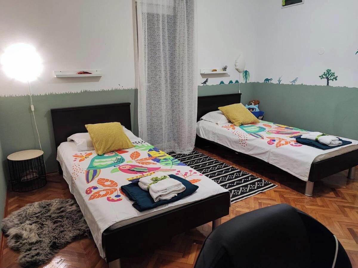 A room with two beds, a round table, a chair, a window with a sheer curtain, and a wall with a nature-themed mural.