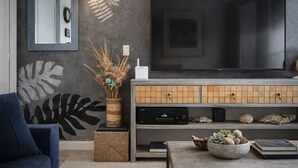 A modern living room with a large flat-screen TV, a dark grey textured wall, a decorative plant, and a blue armchair. - Cabo Oasis Escape - 1 bedroom Apartment (Cabo San Lucas)