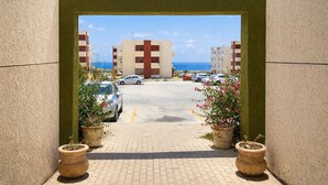 A view through a doorway to a parking area with several cars, potted plants, and a building with a red and white facade in the background. - Cabo Oasis Escape - 1 bedroom Apartment (Cabo San Lucas)