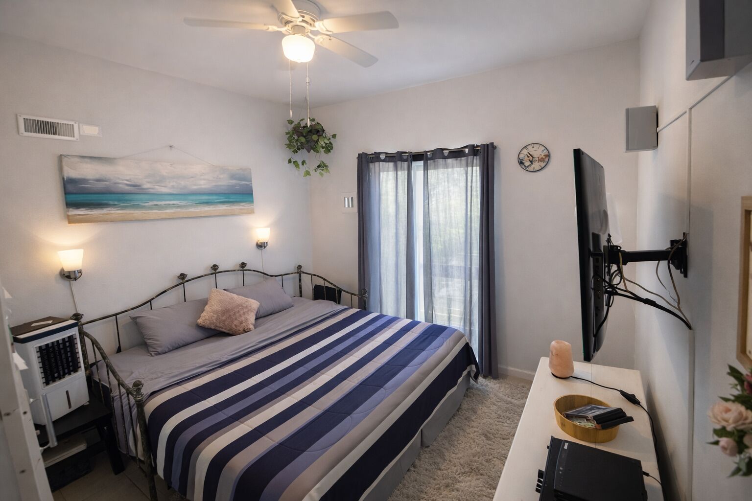 Un dormitorio con cama, ventilador de techo, un cuadro, ventana con cortinas, televisor montado en la pared y escritorio con computadora y planta.